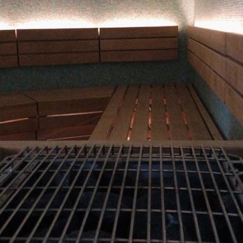 Spa Light - Jade room sauna - The image shows the interior of a sauna room with wooden benches arranged along the walls. The benches are made of horizontal wooden slats. In the foreground, there is a metal grate covering a heater or stove, which is typical in sauna settings. The walls are covered with a textured material, and there is a small vent or heater opening on the wall. The lighting is subdued, creating a calm atmosphere. - Massage Near Me - Lincoln, Rhode Island