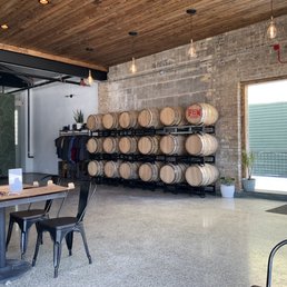 Spacious taproom. - The image shows the interior of a brewery or taproom with a wooden ceiling and brick wall. Multiple wine barrels are stored on black racks along the wall, with some barrels labeled 'FEM.' There are tables with black chairs, and a large window allows natural light into the space. Potted plants are placed near the window, and decorative hanging light bulbs are suspended from the ceiling, contributing to a modern, industrial aesthetic.