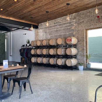 Harbor Brewing - Spacious taproom. - The image shows the interior of a brewery or taproom with a wooden ceiling and brick wall. Multiple wine barrels are stored on black racks along the wall, with some barrels labeled 'FEM.' There are tables with black chairs, and a large window allows natural light into the space. Potted plants are placed near the window, and decorative hanging light bulbs are suspended from the ceiling, contributing to a modern, industrial aesthetic. - Breweries Near Me - Lake Villa, Illinois