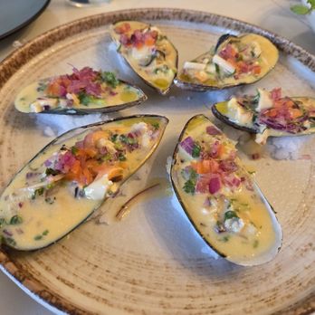 Fusionista - A plate of cooked clams topped with a creamy sauce, chopped herbs, and vegetables, presented on a ceramic dish. - Peruvian Near Me - Montclair, New Jersey