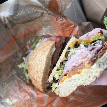 Schlotzsky's - The Original Sandwich - A sandwich featuring sliced deli meats, shredded lettuce, cheese, and black olives inside a sesame seed bun. The sandwich is held in a person's hand, with the background showing part of a car interior and a paper wrapper from a sandwich shop. - Sandwiches Near Me - Appleton, Wisconsin