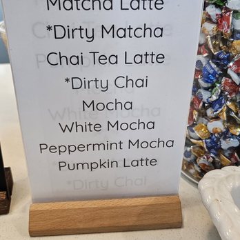 Uptown Cafe - Menu as of April 2024 - A printed menu sign on a wooden stand lists beverage options including Matcha Latte, Dirty Matcha, Chai Tea Latte, Dirty Chai, Mocha, White Mocha, Peppermint Mocha, and Pumpkin Latte. The sign is placed on a counter with a blurred background showing cafe supplies and a container filled with wrapped candies. The menu features a mix of coffee, tea, and specialty drinks. - Coffee & Tea Near Me - Chantilly , Virginia