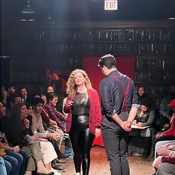 Drunk Shakespeare - Chicago - A woman and a man are on stage in front of an audience, with the woman speaking or singing and the man listening, in a theater with bookshelves and an exit sign overhead. - Performing Arts Near Me - Chicago, Illinois