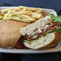 Grilled Chicken BLT - A takeout meal features a chicken salad sandwich with lettuce and tomato on a hoagie bun, accompanied by a portion of French fries. The sandwich is partially cut, revealing the chicken salad filling, and the fries are thin-cut and lightly seasoned. The meal is presented in a rectangular, disposable container, likely for quick service or takeout.
