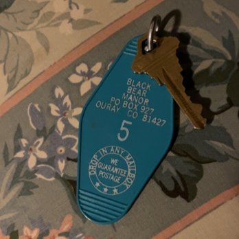 Black Bear Manor - My cute little room key - A blue plastic hotel key tag with the number 5 and address details for Black Bear Manor in Ouray, Colorado, is attached to a brass key. The key and tag are placed on a floral-patterned tablecloth featuring various flowers and leaves in muted colors. The key tag includes a guarantee of postage and the address information for the hotel. - Bed & Breakfast Near Me - Ouray, Colorado