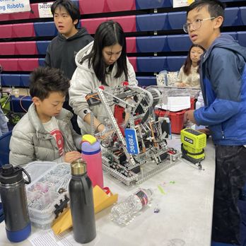 Rolling Robots - Irvine - Several children are engaged in working on a robotics project at a table, with one girl adjusting the robot and others observing. The table has water bottles, a toolbox, and tools. The background features colorful tiered seating and signs, indicating an indoor event or competition related to robotics or STEM activities. - Summer Camps Near Me - Irvine, California
