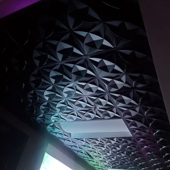 Vybe - Ceiling - Ceiling displays a repeating pattern of circular, petal-like shapes arranged in a geometric design. The room is dimly lit, with subtle colorful lighting casting hues of purple, blue, and green on the ceiling. A rectangular light fixture is embedded in the ceiling, and part of a window or screen is visible in the lower left corner, providing some natural or external light. - Venues & Event Spaces Near Me - Chandler, Arizona