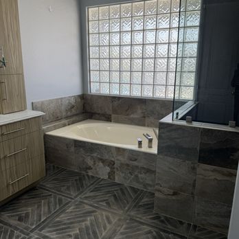 Marshall Renovations - Stunning spa area in primary bathroom - The bathroom includes a built-in bathtub surrounded by gray stone tiles, positioned beneath a large glass block window that provides natural light. To the left, there is a wooden cabinet with multiple drawers and a marble countertop. The floor is covered with dark, patterned tiles arranged in a diagonal herringbone pattern, and a glass shower enclosure is partially visible on the right side of the image. - General Contractors Near Me