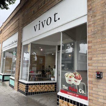Vivot Craft Cafe - Outside - The image shows the exterior of Vivot, a café located in a brick building. The storefront features large glass windows, allowing a view of the interior where patrons are seated. A white sign above the window displays the name "vivot" in lowercase black letters. There is also a promotional poster on the window advertising "The Poke Co." with images of poke bowls and the slogan "SHAKE IT UP!". The sidewalk in front is concrete, and part of a tree is visible in the upper left corner. - Bubble Tea Near Me - Fullerton, California