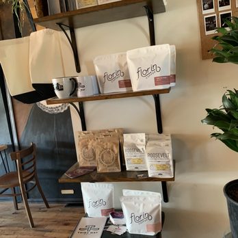 Third Way Cafe - A wooden shelf with black metal supports holds various coffee products, including bags labeled 'Florin Coffee' and 'Roosevelt Coffee Roasters,' along with a ceramic mug and a small container. Below, a small table displays additional coffee bags and promotional materials. The background includes a corkboard with photos and a potted plant, indicating a cozy coffee shop environment. - Coffee & Tea Near Me - Columbus, Ohio