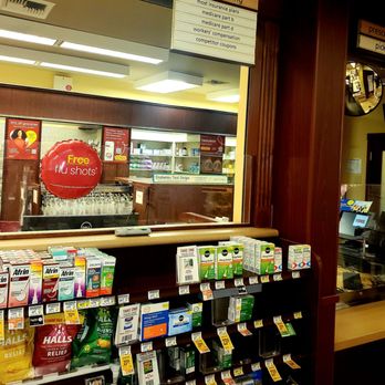 Vons Pharmacy - Pharmacy - Pharmacy shelf displaying various allergy relief and flu medication boxes at Vons Pharmacy in Pasadena, California. The shelf includes products like Afrin, Zicam, and Hall's cough drops, with price tags and promotional signs. Behind the shelf, a window shows the pharmacy interior with a sign advertising free flu shots and a list of accepted insurance plans. The setting features wood paneling and a counter area with a computer and pharmacy supplies. - Pharmacy Near Me - Pasadena, California