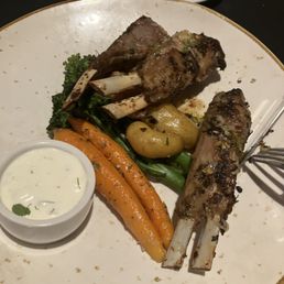 Rack of lamb - A white plate holds grilled lamb chops with visible grill marks, accompanied by roasted potatoes, carrots, broccoli, and green beans. A small bowl of creamy herb sauce is placed on the side. The dish is presented on a dark table, with a smaller empty plate visible in the background.