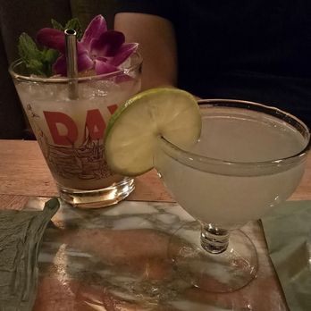 Daydream - Mai tai & classic daquri - The image shows two cocktails on a wooden table in a dimly lit environment. One drink is in a coupe glass with a lemon slice garnish, and the other is in a glass with floral and mint decorations, featuring a straw. A person wearing a black shirt is partially visible in the background. - Cocktail Bars Near Me - Albuquerque, New Mexico