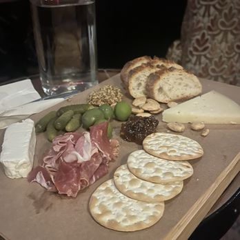 J9 Wine Bar - Charcuterie board - A wooden tray holds a charcuterie assortment including bread slices, cheese, green olives, prosciutto, and spreads, with a glass of water nearby. - Wine Bars Near Me - Chicago, Illinois