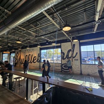 Pins Mechanical - Several individuals are engaged in shuffleboard games inside a spacious, industrial-themed venue with large windows and exposed ceiling pipes. - Bowling Near Me - Austin, Texas