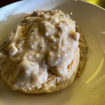 Lemon Leaf Cafe - 1/2 order of biscuits & gravy - A biscuit topped with sausage gravy is placed in a white bowl. The gravy appears creamy with visible bits of sausage. The image is taken indoors, with natural lighting highlighting the dish. The setting suggests a breakfast or brunch meal at Lemon Leaf Cafe in Largo, FL. - Breakfast & Brunch Near Me - Largo, Florida