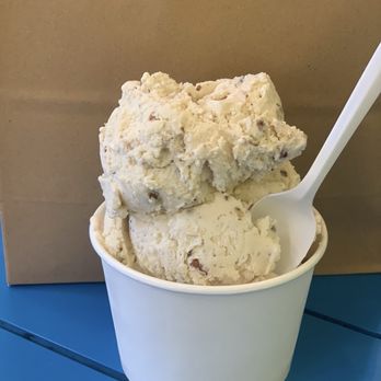 Granite State Candy Shoppe - Large NH Maple Pecan - A cup of ice cream with visible mix-ins and a white plastic spoon, placed on a blue surface with a plain background. - Candy Stores Near Me - Concord, New Hampshire