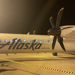 Smaller air craft - A small aircraft with Alaska Airlines branding, including the logo and the word 'Horizon,' is parked on the airport tarmac during nighttime. The airplane features a propeller engine with four blades, and the scene is illuminated by airport lighting. The background shows a dark sky, and the ground has yellow and black striped markings for safety and guidance.
