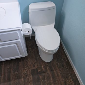 Drain Control - A white toilet is situated beside a small white cabinet with a roll of toilet paper hanging on the side, in a bathroom with light blue walls and dark wood flooring. - Plumbing Near Me - Richmond, Texas