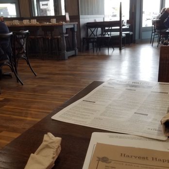 First Watch - Cute space - The image shows the interior of a restaurant with wooden floors and a mix of tables and chairs. Large windows allow natural light to fill the space, and pendant lights hang from the ceiling. There are some patrons seated, and the decor includes a partition wall with shelves. The foreground features a table with menus and a wrapped utensil. The overall atmosphere appears casual and inviting. - Breakfast & Brunch Near Me - Mequon, Wisconsin