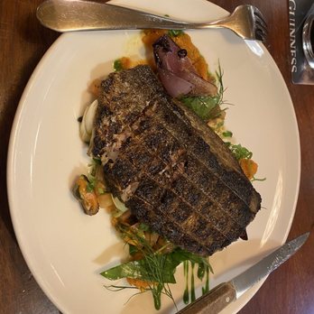 Spitalfields - A grilled fish fillet with crispy skin is presented on a white plate, accompanied by vegetables and garnishes, on a wooden table. - Pubs Near Me - Dublin 8, D