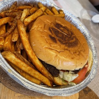 Pantano's Deli Plainview - The Cajun Burger - A meal featuring a burger with a toasted bun, lettuce, tomato, and a beef patty, served in a disposable foil container. The burger bun has a slightly charred top. Alongside the burger, there are seasoned French fries, some with a reddish spice coating. The meal is placed on a wooden board, with a paper napkin and condiment packet visible nearby, suggesting a casual dining setting. - Italian Near Me - Plainview, New York