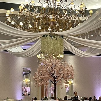 Meridian Banquets - The image shows a banquet hall decorated for an event, with a large ornate chandelier hanging from the ceiling. White fabric is draped across the ceiling in a decorative manner. A pink cherry blossom tree centerpiece is positioned in the middle of the room, surrounded by tables with tablecloths and place settings. The walls have mirrors and soft lighting, and there are guests and staff present, indicating a formal gathering or celebration. - Venues & Event Spaces Near Me - Rolling Meadows, Illinois