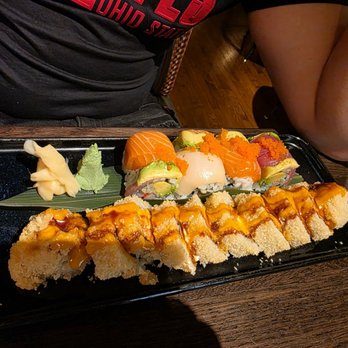 The 698 - Rainbow  Roll (back) Sunshine roll (front) - A black rectangular tray holds an assortment of sushi rolls, including a roll topped with eel sauce, and others with fish, avocado, and roe. Wasabi and pickled ginger are placed on the side. The sushi is presented on a green leaf, with a wooden table surface visible beneath. The setting appears to be a restaurant with dim lighting and wooden flooring. - Sushi Bars Near Me - Columbus, Ohio