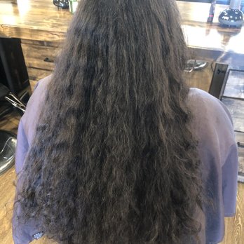 Salon Estique - Before hair cut - A person with long, dark, curly hair is seated in a salon chair, facing away from the camera, in a salon setting with a wooden counter and hair care products. - Hair Salons Near Me - Phoenix, Arizona