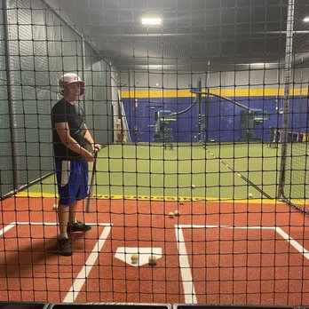 Humdingers - A person in a batting cage wearing a helmet, black shirt, and blue shorts, holding a baseball bat, with baseballs on the ground. - Arcades Near Me - Paramus, New Jersey