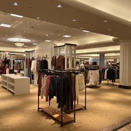 The image shows the interior of a retail clothing store with multiple clothing racks filled with various garments. The store features warm, ambient lighting from ceiling fixtures, including a central ceiling light. Mannequins and display shelves are visible, and the space is spacious with a beige carpeted floor and white columns. The store appears well-organized, with sections for different clothing styles and categories.
