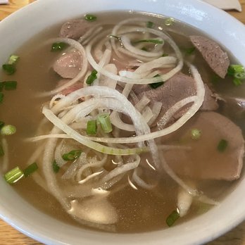 Pho Zabb - A bowl of Vietnamese pho contains sliced beef, thinly sliced onions, and chopped green onions in a clear, light broth. The dish is served in a white ceramic bowl placed on a wooden table, with lime wedges partially visible on the side. The ingredients are arranged with the beef slices submerged and the onions and green onions floating on top, creating a traditional presentation of Vietnamese noodle soup. - Asian Fusion Near Me - Boston, Massachusetts
