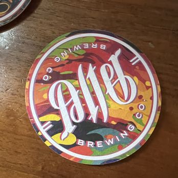 Dan's Pizza - Coaster - A circular coaster displaying the Half Acre Brewing Co. logo with a colorful, abstract background. The coaster is placed on a wooden table surface, and part of another similar coaster is visible in the top left corner. The design includes the brewery's name and branding elements, with a vivid, multicolored pattern. - Pizza Near Me - Downers Grove, Illinois