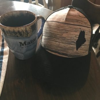 Katahdin General Store - A coffee mug I got a few years ago and the hat-I LOVE! Just got this last week!! - A ceramic coffee mug with a Maine-themed design, including the state outline, is placed on a dark wooden table. Next to it is a small, rounded backpack with a wood grain pattern and a black Maine silhouette. The background shows a chair and part of a room with natural light coming through a window. - Convenience Stores Near Me - Millinocket, Maine