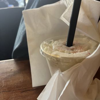 The Burger Social - Peppermint milkshake to go! SO GOOD - A peppermint milkshake in a clear plastic cup with a black straw is wrapped in white paper. The drink appears to have whipped cream and peppermint pieces on top. The cup is on a wooden table, with a blurred background showing a window and some outdoor scenery. - Bars Near Me - Wheaton, Illinois