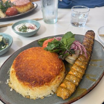 Joon - A plate of Persian cuisine includes a mound of saffron-infused rice, a grilled kebab, and fresh herbs and vegetables. The rice has a crispy golden crust on top, and the kebab is seasoned and grilled. Garnishes include parsley, tomato, and onion slices. The meal is served on a ceramic plate, with additional dishes and drinks visible in the background, indicating a restaurant setting. - Persian/Iranian Near Me - Vienna, Virginia