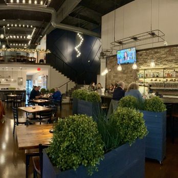 Barley Thyme Kitchen Plus Brew - Inside of restaurant - The image shows the interior of a modern restaurant with a high ceiling and contemporary decor. There are several dining tables with chairs, some occupied by patrons. The bar area has multiple TVs mounted above it, and a brick wall behind the bar. A staircase leads to a second-floor balcony with additional seating. Potted plants are placed in the foreground, and the lighting includes hanging bulbs and string lights, creating a warm atmosphere. - Brewpubs Near Me - Danville, Indiana