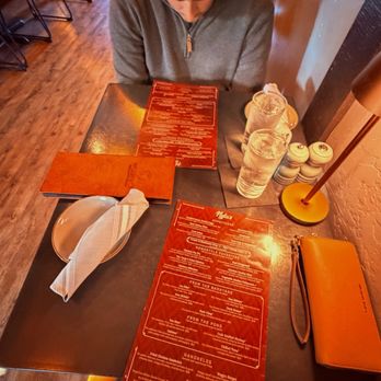 Nyla's Restaurant - Two person table upstairs - A person with dark hair tied back, wearing a gray zip-up sweater, is seated at a dark-colored table in a restaurant. The table has a red menu, two glasses of water, a napkin, and salt and pepper shakers. The restaurant features wooden flooring and a warm, ambient lighting. The setting appears casual and intimate, with additional seating visible in the background. - Cocktail Bars Near Me - Westfield, Indiana
