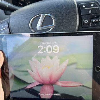 CompuTech - Ipad was fixed - A person is holding a tablet with a flower wallpaper and the time 2:09 displayed on the screen, inside a Lexus vehicle. The dashboard and steering wheel with the Lexus logo are visible in the background. The tablet screen also shows the date, Friday, October 10, and a battery indicator at 67%. The interior of the car includes controls on the steering wheel and instrument gauges behind it. - IT Services & Computer Repair Near Me - Garden Grove, California