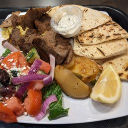 Greek Salad - A rectangular white plate contains grilled pita bread, slices of grilled meat, roasted potatoes, and a lemon wedge. A small container of sauce is placed on the plate. A side salad with chopped tomatoes, cucumbers, red onions, black olives, and feta cheese is also present. The plate is on a black tray, and the food appears freshly prepared.