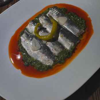 Asador Bastian - Sardine - The image shows a dish featuring sardines placed on a green sauce, possibly made from herbs or vegetables. The sardines are garnished with a yellow pepper and a small piece of garlic. The dish is presented on a white, oval-shaped plate with a border of red oil around the green sauce, creating a colorful contrast. The presentation emphasizes the seafood and vibrant garnishes. - Spanish Near Me - Chicago, Illinois