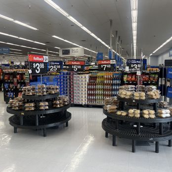 Walmart Supercenter - a variety of products on display - Inside a Walmart Supercenter, baked goods such as cookies are displayed on black circular shelves. The store has bright lighting, white tiled floors, and overhead signage indicating product prices and discounts. The background shows aisles with clothing and other merchandise, with a few shoppers visible. - Department Stores Near Me - Willows, California