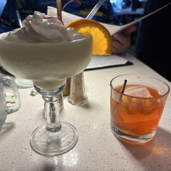Tiesto's Restaurant - Frozen Pina Colada, Negroni - A frozen piña colada served in a glass with whipped cream, a cherry, and an orange slice garnish, is placed on a table. Next to it is an orange-colored cocktail with ice cubes and lemon slices in a short glass. The table surface is light-colored, and there are other objects and people in the background. - Latin American Near Me - Patchogue, New York