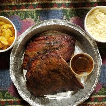 Mr E Chefs Barbecue - The image shows two large barbecue ribs with a dark, seasoned crust placed on a round metal tray. Two small bowls contain side dishes: one with potato casserole topped with melted cheese and the other with potato salad. The tray is set on a colorful, patterned fabric surface. - Caterers Near Me - Chicago, Illinois
