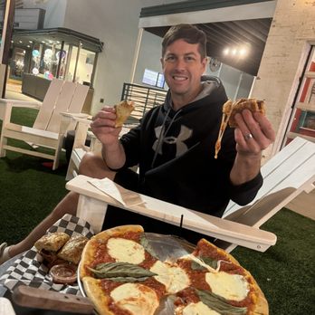 The Station - A man is sitting outdoors at a restaurant, holding a slice of pizza in each hand and smiling at the camera. He is wearing a black hoodie with a logo and has short dark hair. In front of him, there is a tray with a partially eaten pizza topped with cheese, tomato sauce, and basil leaves. The background features string lights hanging overhead, a white brick wall, and outdoor furniture, indicating a casual dining setting with a relaxed ambiance. - Pizza Near Me - Jackson, Mississippi