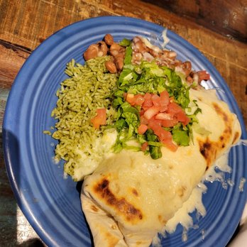 Range Cafe - Bean burrito with green rice. - The image shows a blue ceramic plate containing a burrito covered with melted cheese and garnished with chopped tomatoes and lettuce. Accompanying the burrito are green rice, refried beans, and a small portion of beans with sauce. The plate is placed on a wooden table, with part of another dish visible in the background. The setting appears to be a casual dining restaurant. - Breakfast & Brunch Near Me - Albuquerque, New Mexico