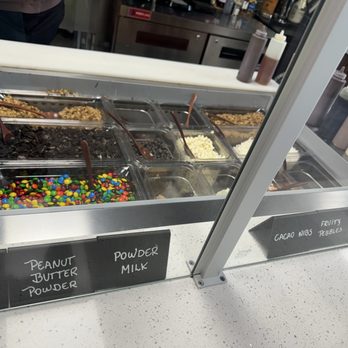 Suko Cafe - More toppings - A dessert topping station with multiple containers holding candies, nuts, and powders, labeled with handwritten signs. - Acai Bowls Near Me - North Reading, Massachusetts