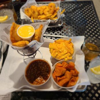 Backyard Barbeque - 3 cheese Collard green Empanada, Baked Beans, Smoked Mac N Cheese - A food tray with fried fish, mashed sweet potatoes, baked beans, and drinks on a black metal table, with people in the background. - Barbeque Near Me - Freeport, New York