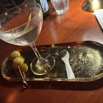 CODE RED - A cocktail glass containing a clear drink with a lemon twist garnish is placed on a gold tray. The tray also holds three green olives on a skewer and a small spoon with black caviar. The setting appears to be in a dimly lit environment, likely a bar or restaurant, with a wooden table surface and some additional items like a glass of water, a menu, and a lamp in the background. - Cocktail Bars Near Me - Washington, DC, District of Columbia