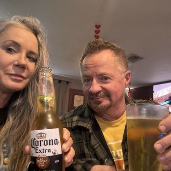 Butts and Buns - A woman and a man are posing together indoors, each holding a drink. The woman holds a Corona beer bottle with lime, and the man holds a glass of beer. The background includes a television and Valentine's Day decorations, such as small red hearts hanging from the ceiling. The setting appears to be a casual social gathering or restaurant. - Food Trucks Near Me - Payson, Arizona
